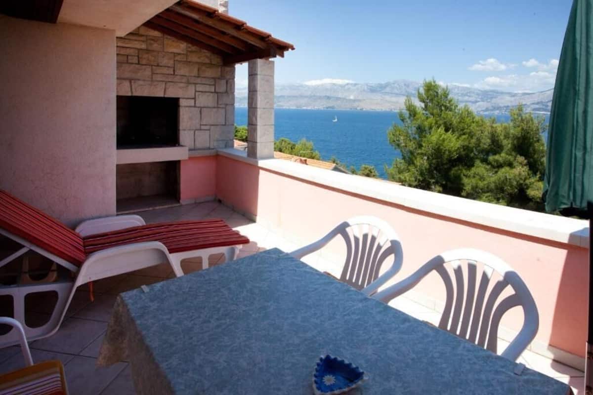 A balcony with a stone fireplace, white plastic chairs, a table, and a view of the sea and mountains.