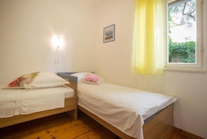 A small, single-room accommodation with two single beds, a bedside table, a wall-mounted light, a window with yellow curtains, and a framed picture on the wall. - Apartments Damir (Postira)