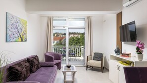 A modern living room with a purple sofa, a small coffee table, a TV on a stand, and a balcony with a view of the city. - Apartments Star 2 (Dubrovnik)