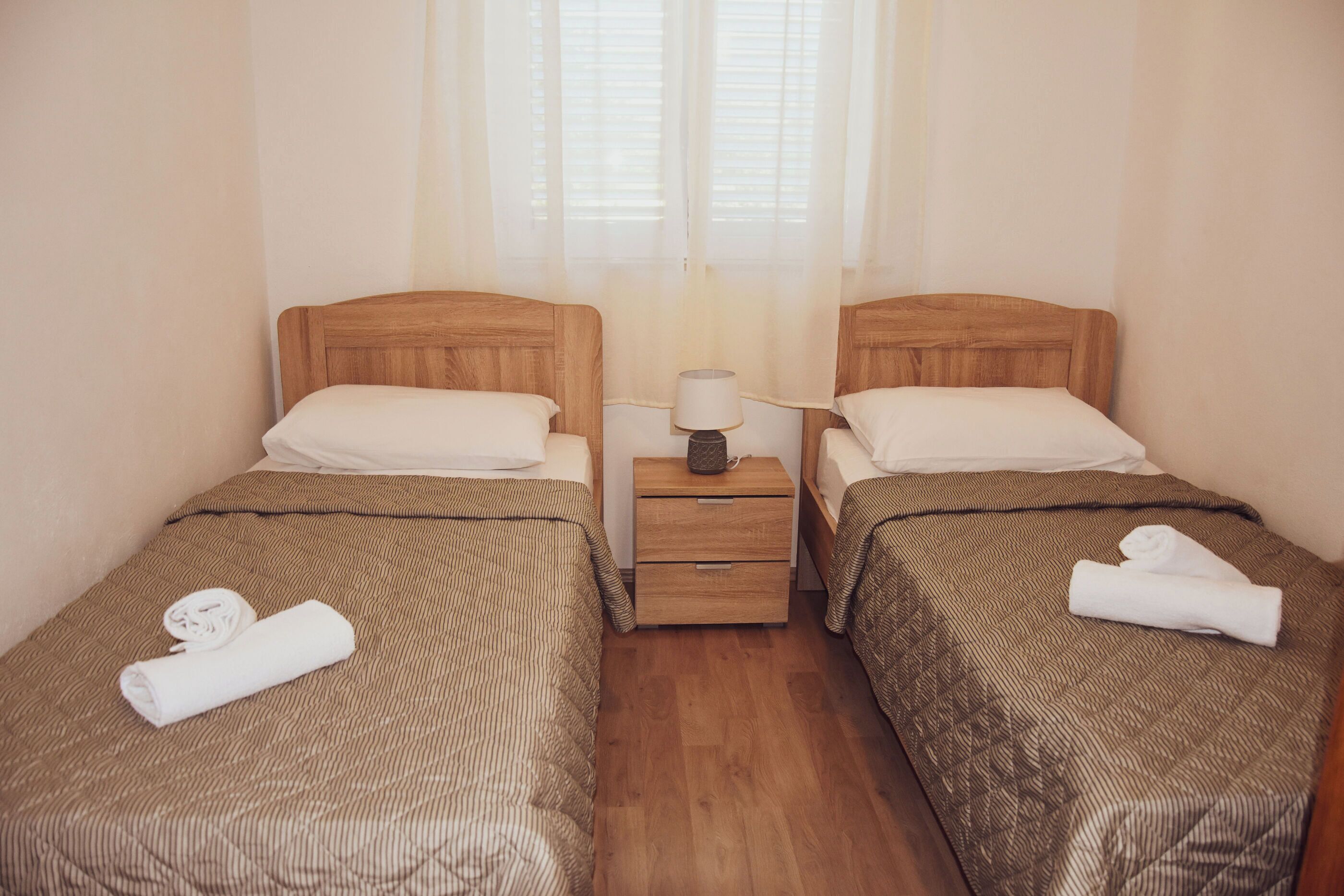 Ein Doppelzimmer mit zwei Betten, Holz-Kopfteile, einem Nachttisch mit Lampe und einem Fenster mit transparenten Vorhängen.