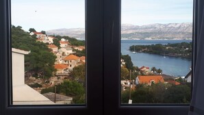 A view through a window of a coastal town with houses, a body of water, and mountains in the background. - Apartment Ivope (Supetar)