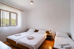A room with two single beds, a wooden floor, a window with sheer curtains, and a bedside table with a lamp. - Apartment Ivope (Supetar)