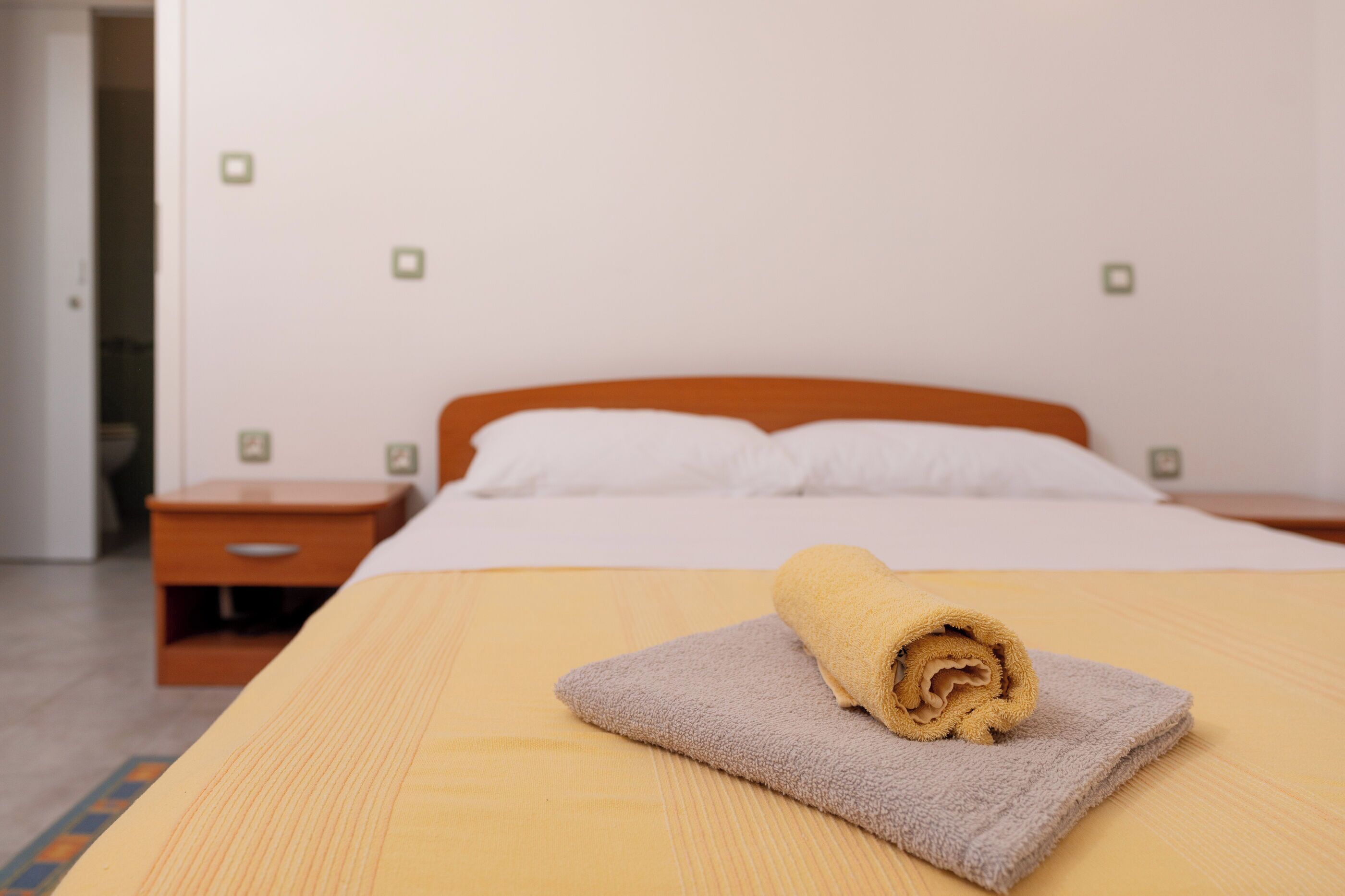 Ein ordentlich bezogenes Bett mit einem zusammengerollten Handtuch auf einer grauen Decke, ein Nachttisch aus Holz mit einer Schublade und eine weiße Wand mit kleinen, quadratischen Dekorationen.