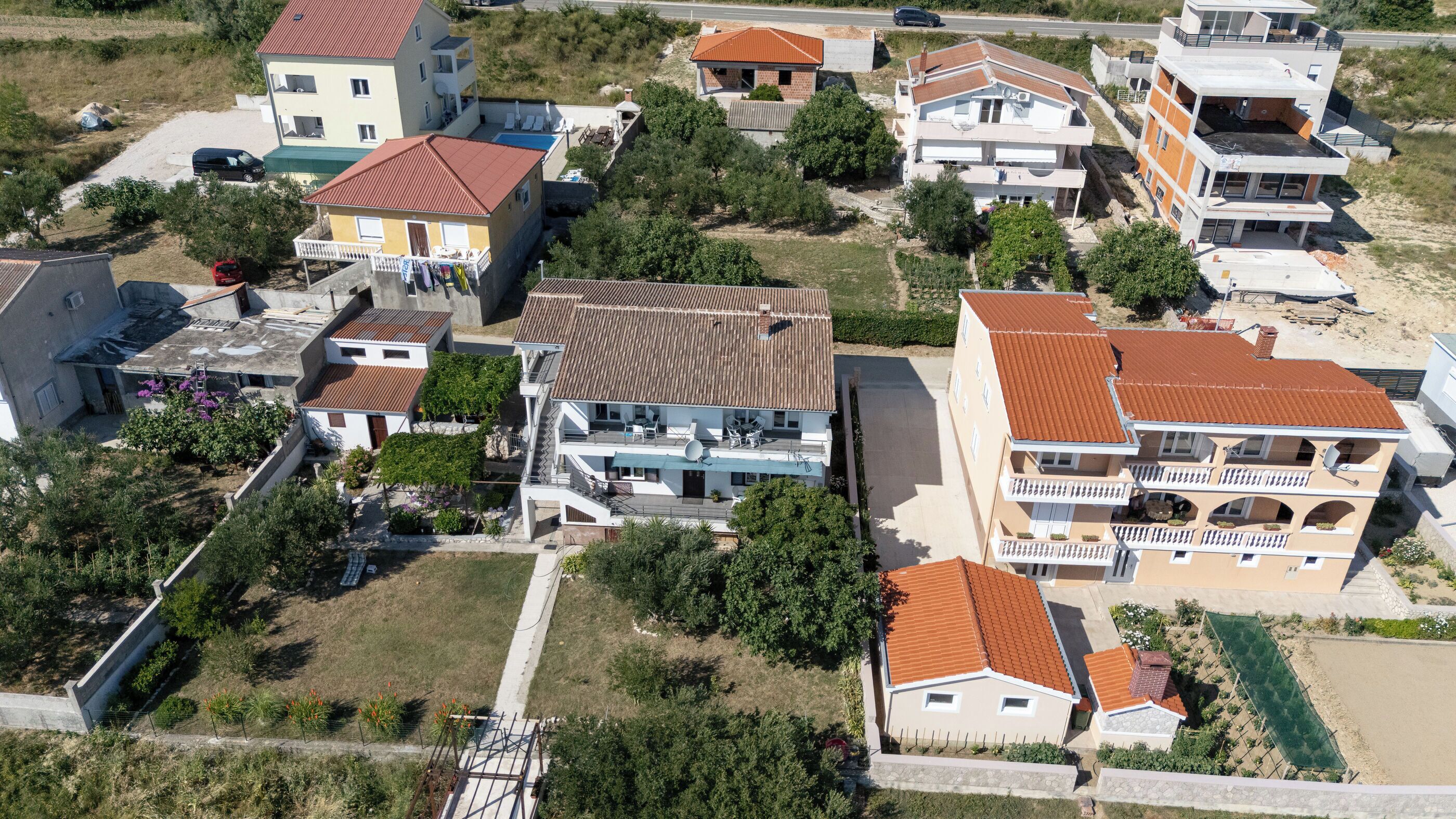 An aerial view of a residential area with multiple houses, some with balconies and satellite dishes, surrounded by greenery and a road.