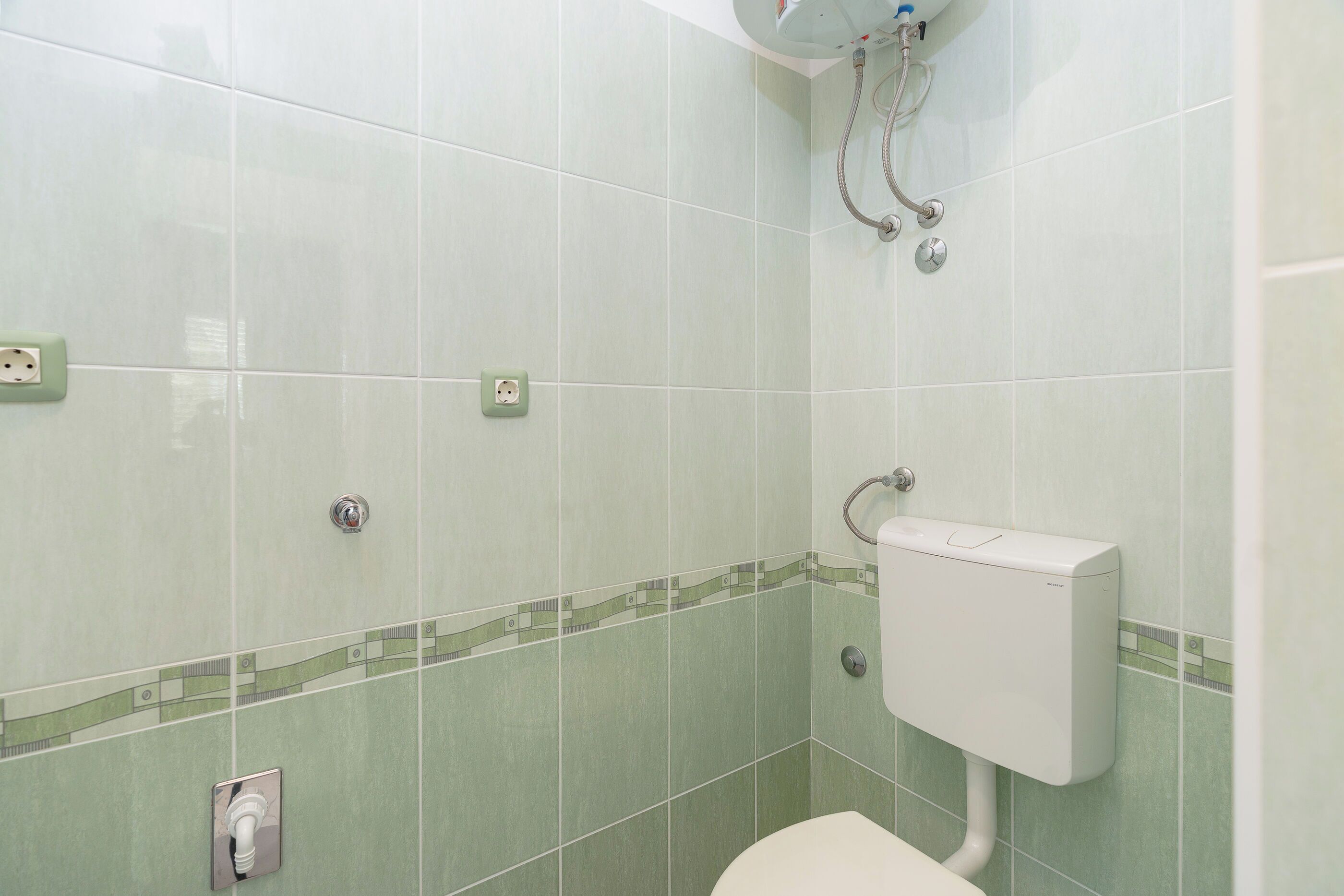 A bathroom with green tiles, a toilet, a shower with a handheld showerhead, and a wall-mounted faucet.
