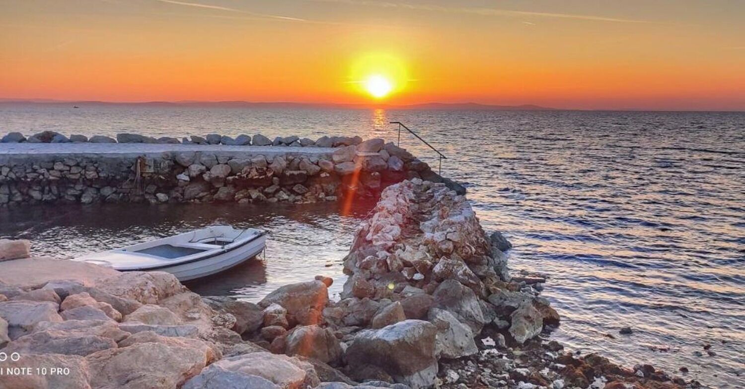 Una costa rocosa con un bote amarrado cerca de un muro de piedra, y el sol poniéndose sobre el agua.