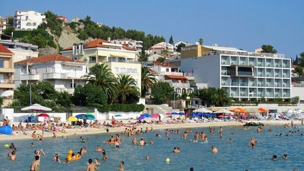 A crowded beach with people swimming and sunbathing, surrounded by buildings and palm trees. - Apartments and Rooms Robi (Podstrana)