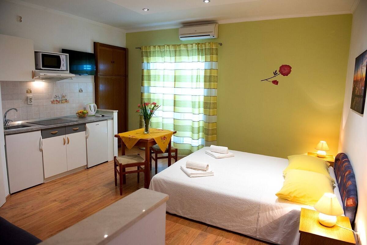A compact hotel room with a kitchenette, a dining area, and a bed with a nightstand.