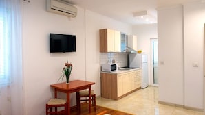 A compact living space with a kitchenette, a dining area with a table and chairs, a flat-screen TV mounted on the wall, and a small window with a curtain. - Apartments and Rooms Robi (Podstrana)