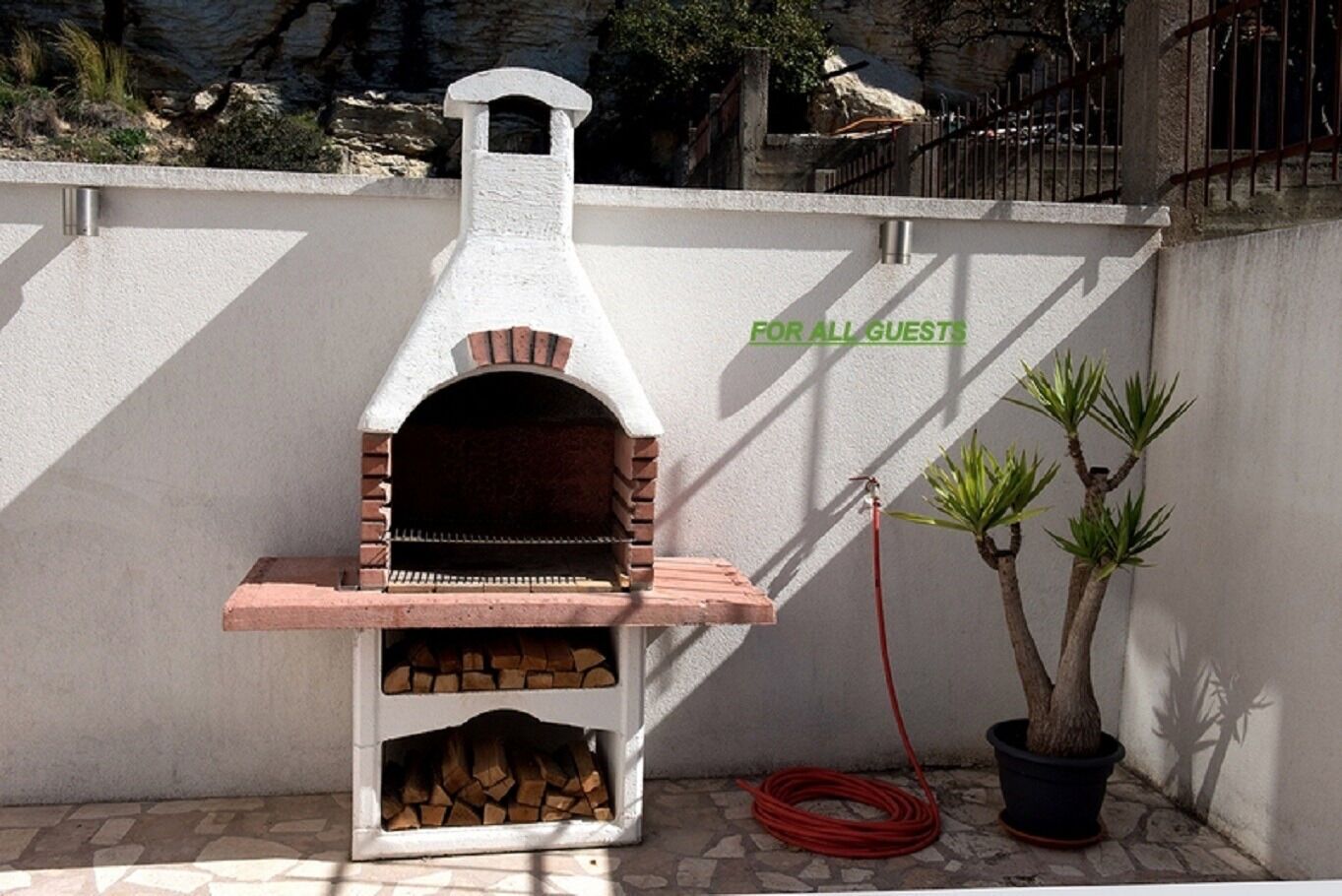 A brick-built outdoor oven labeled "FOR ALL GUESTS" with firewood stored inside and a red hose nearby.