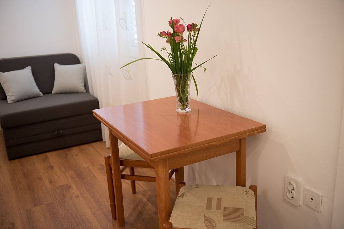 A small wooden table with a vase of flowers, a chair, and a sofa in a room with wooden flooring and white walls.