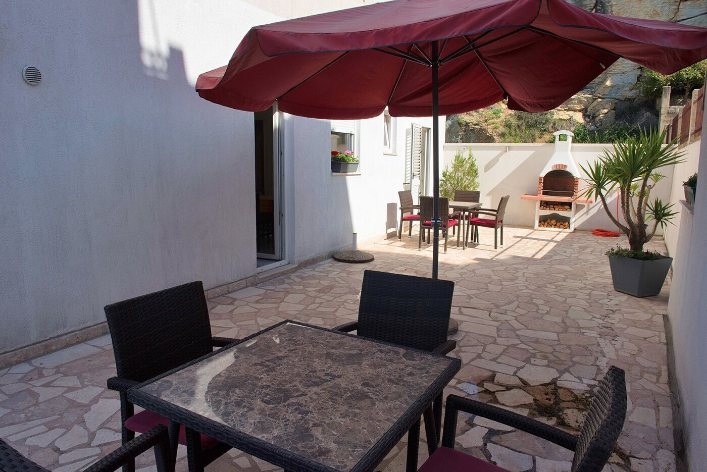 Eine Terrasse im Freien mit Tisch, Stühlen, einem roten Sonnenschirm und einem Grill.