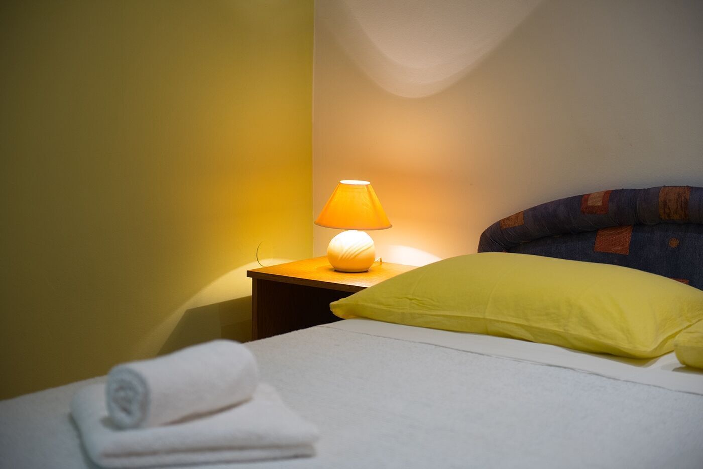 A neatly made bed with a yellow pillow and a white towel, a bedside lamp, and a wooden nightstand.