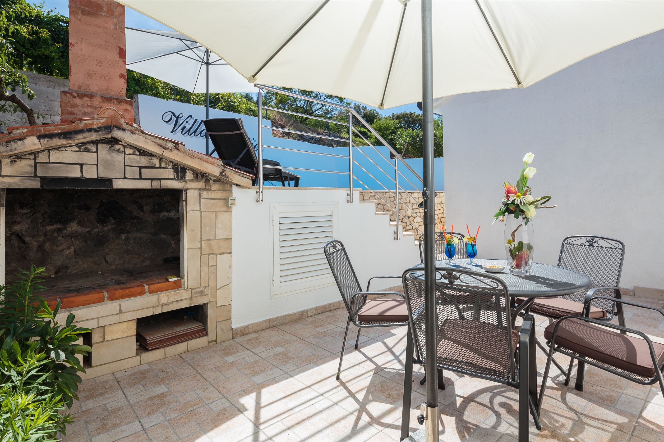 An outdoor patio with a stone fireplace, metal chairs, a table, and a large umbrella providing shade.