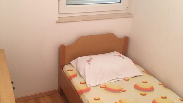 Ein kleines Zimmer mit einem Einzelbett, einem Holzkopfteil, einem Fenster mit Jalousien und einer gestreiften Bettdecke.