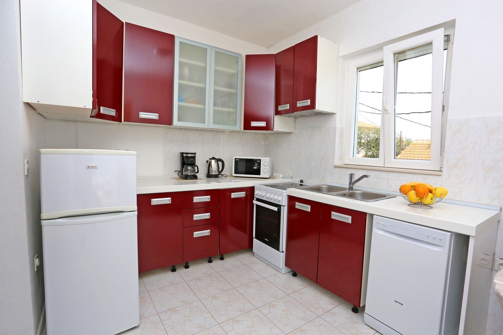 Una cocina compacta con armarios rojos, un refrigerador blanco, microondas, cafetera y lavavajillas.