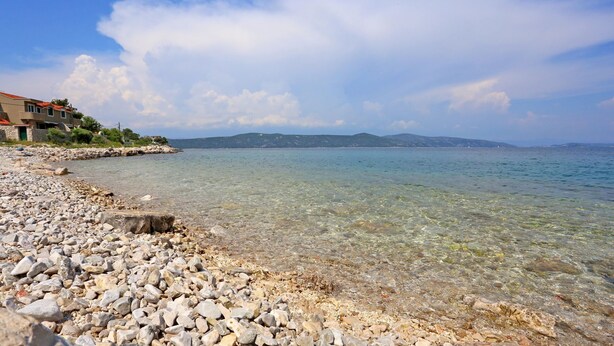 A rocky beach with clear water and a distant view of hills. - Apartment Marija (Trogir)