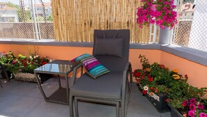 Un balcon avec une chaise en osier, une petite table et un coussin. De part et d’autre, on trouve des plantes en pot, certaines fleuries.