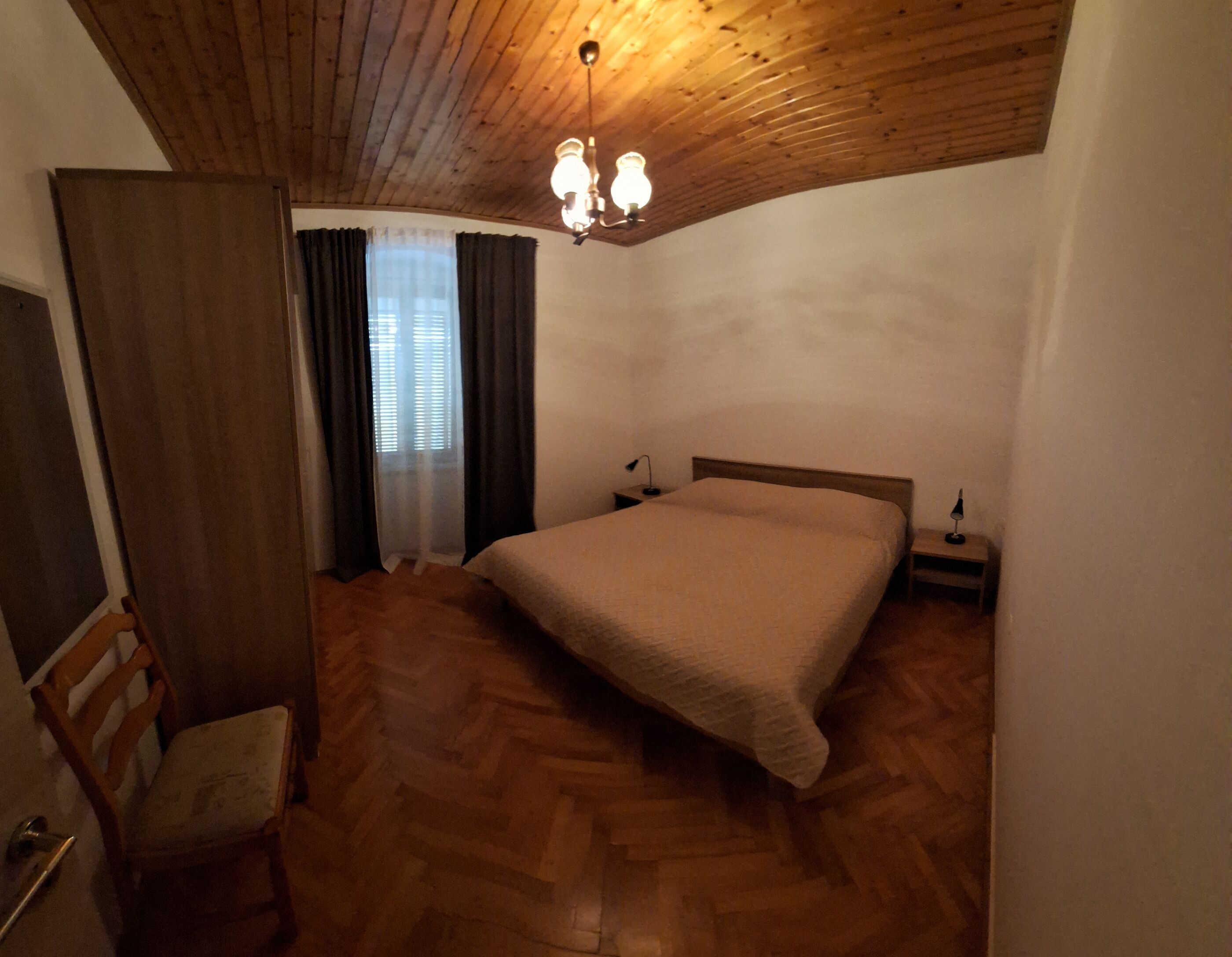 Un dormitorio con techo de madera, una cama, una mesita de noche, una silla y una ventana con cortinas.