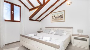 Een slaapkamer met een groot bed, twee nachtkastjes, een plafond met houten balken, een lijst met een afbeelding aan de muur en een raam met gordijnen.