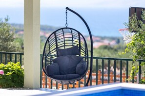 A hanging chair with cushions by a poolside, a metal railing, and a view of the sea. - House Mario (Supetar)