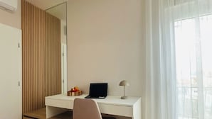Une chambre moderne avec un bureau blanc, un ordinateur portable, une lampe, une chaise, un miroir et une fenêtre ornée de rideaux légers.