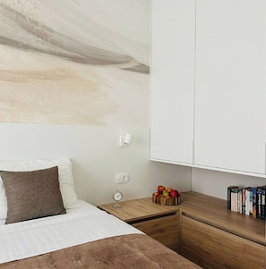 Un dormitorio con una cama, una mesita de noche, una mesita auxiliar de madera, una lámpara de pared, un reloj y una estantería.