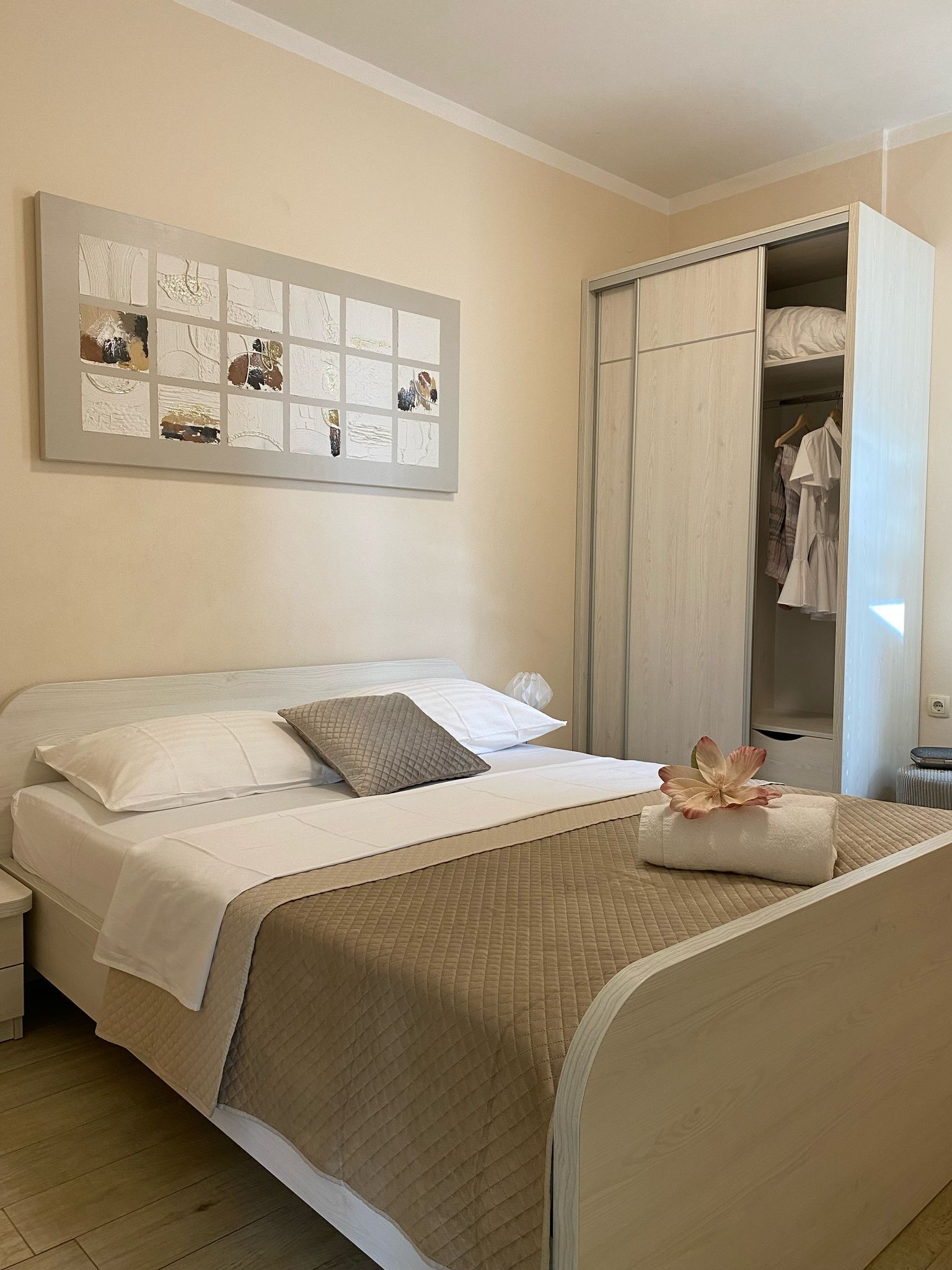 A neatly made bed with a quilted bedspread and pillows, a wall-mounted artwork, and a wardrobe with hanging clothes.