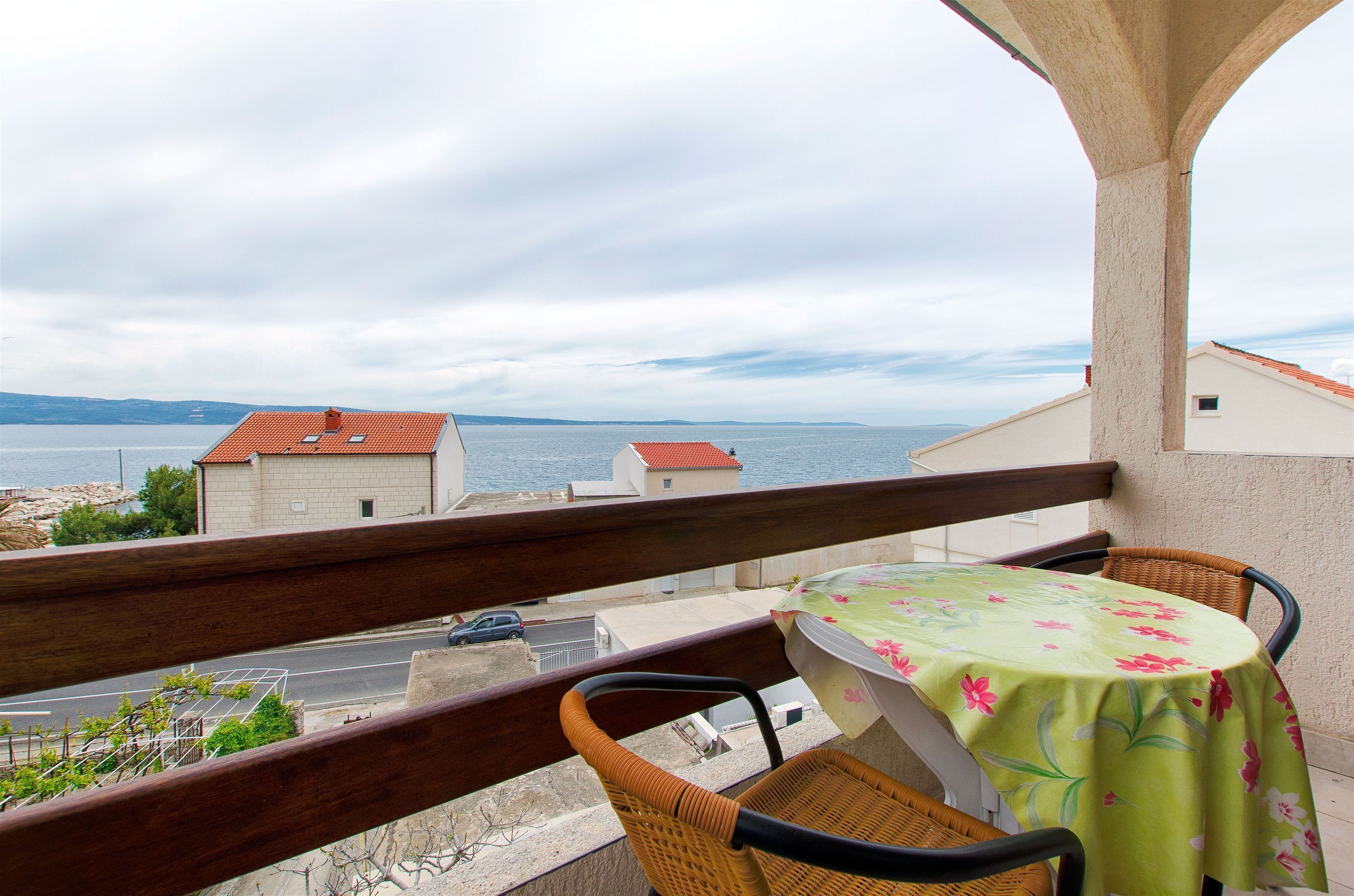 A balcony with wicker chairs and a table, overlooking a coastal view with buildings and a road.