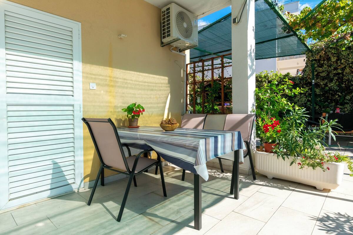 Eine Terrasse mit Tisch, Stühlen, einer Topfpflanze und einer weißen Wand mit einem Fensterladen.