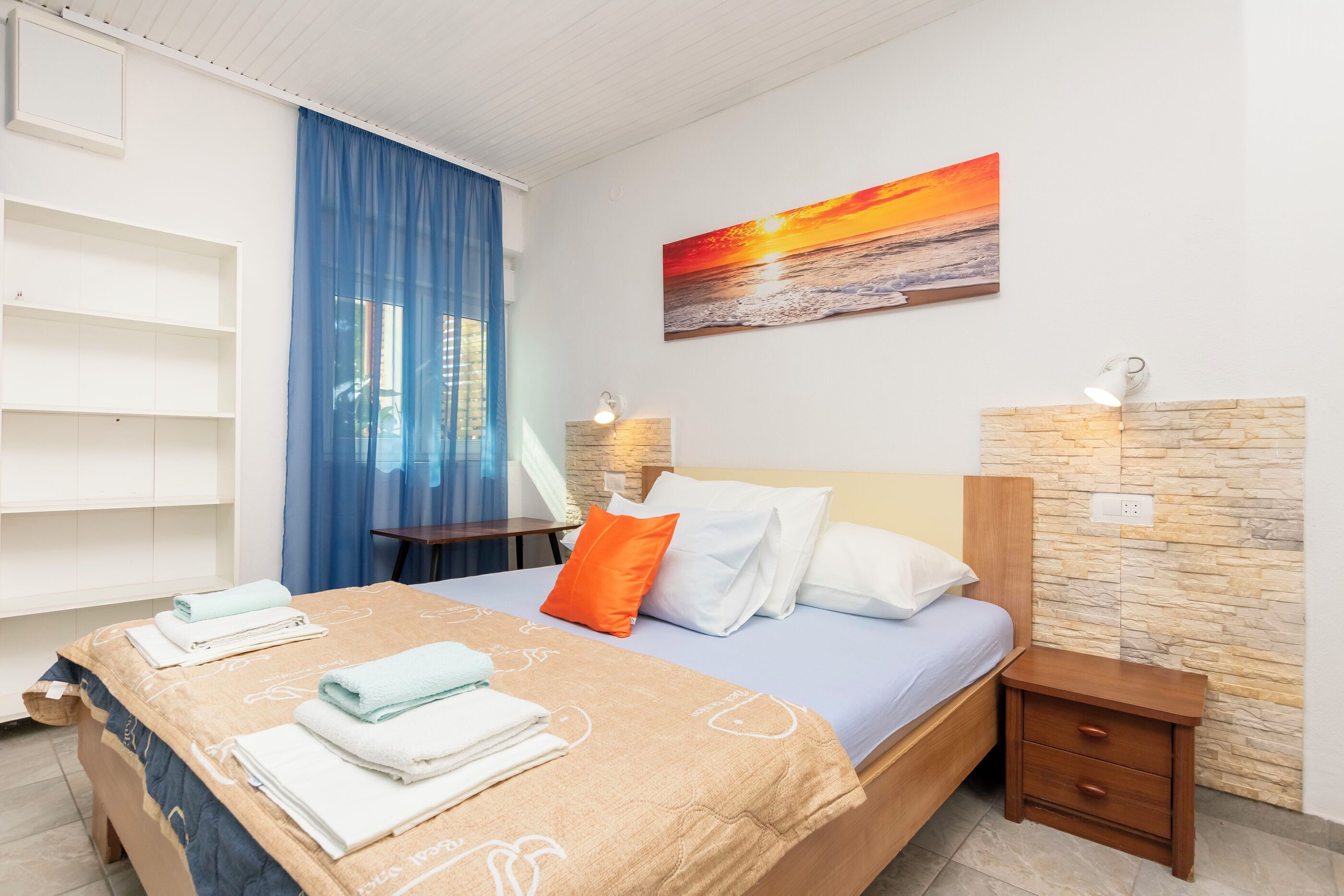 A hotel room with a bed, a nightstand, a wall-mounted shelf, and a painting of a sunset.