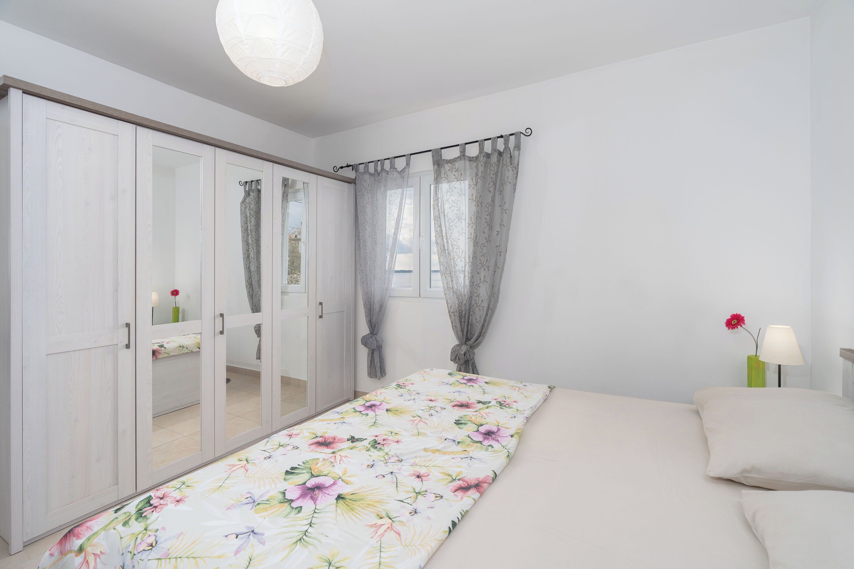 Un dormitorio con cama, armario, ventana con cortinas, mesita de noche con lámpara y un jarrón con una flor.