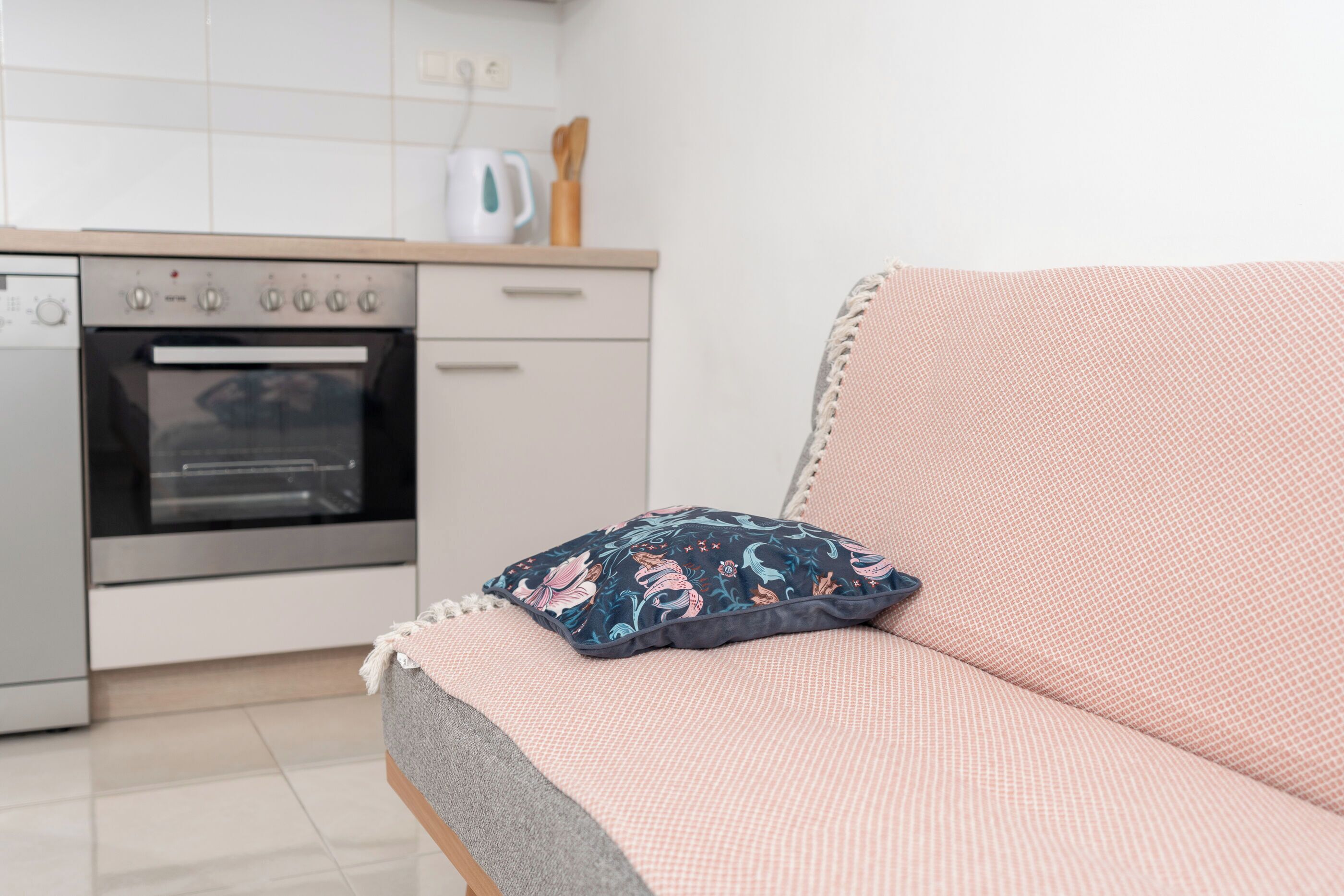 A kitchenette with an oven and a coffee maker, a sofa with a patterned pillow, and a tiled floor.