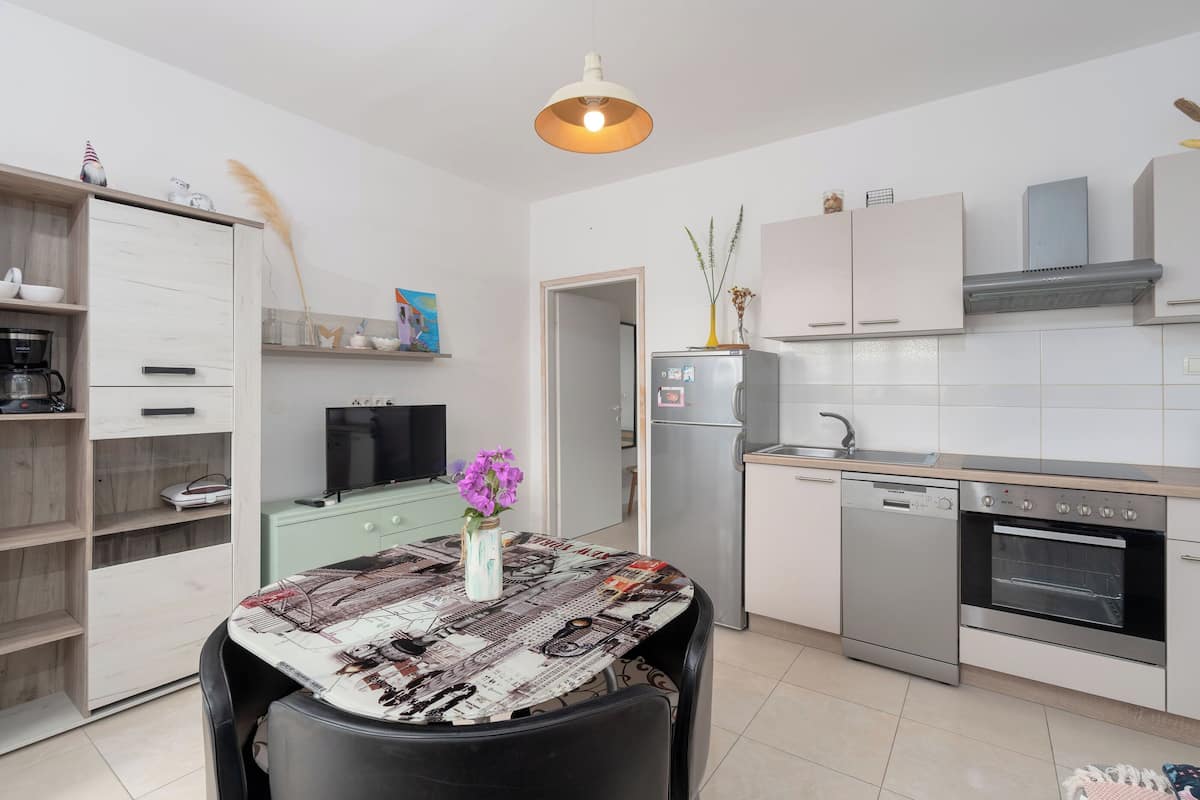 Una cocina compacta con comedor, muebles blancos, lavavajillas, horno y televisor.