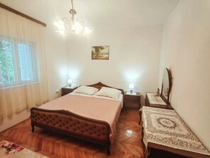 Ein Schlafzimmer mit einem Bett, zwei Nachttischen, einer Chandelüster, einem Fenster mit Vorhängen, einem Gemälde und einem Spiegel.