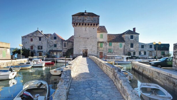 A stone pathway leading to a historic building with a tower, surrounded by water and boats. - Apartment Gordana (Kastela)