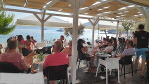 A covered outdoor seating area with tables and chairs, overlooking a beach and the sea. - Apartment Gordana (Kastela)