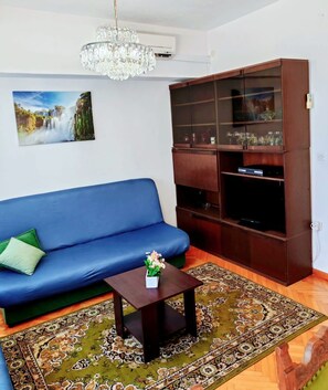Een woonkamer met een blauwe bank, een houten tv-meubel, een kroonluchter en een kamerkleed met een patroon.