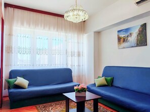 Ein Wohnzimmer mit zwei blauen Sofas, einer Kristallleuchter, einem Gemälde eines Wasserfalls und einem Fenster mit transparenten Vorhängen.