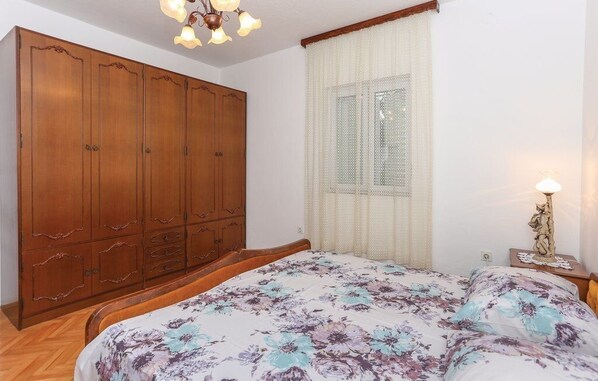 Un dormitorio con un gran armario de madera, una cama con ropa de cama floral, una ventana con cortinas de gasa y una mesita de noche con una lámpara y un adorno.
