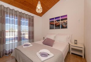 A bedroom with a bed, curtains, a chandelier, a nightstand, and a wall-mounted picture. - House Kristiana (Supetar)