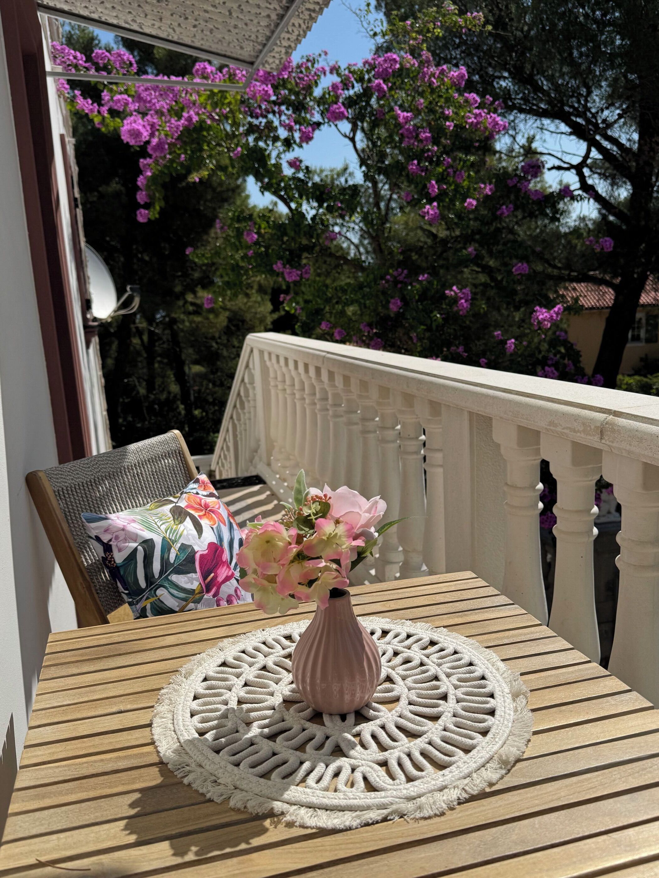 A balcony with a table, a chair, a vase with flowers, and a view of trees with purple flowers.