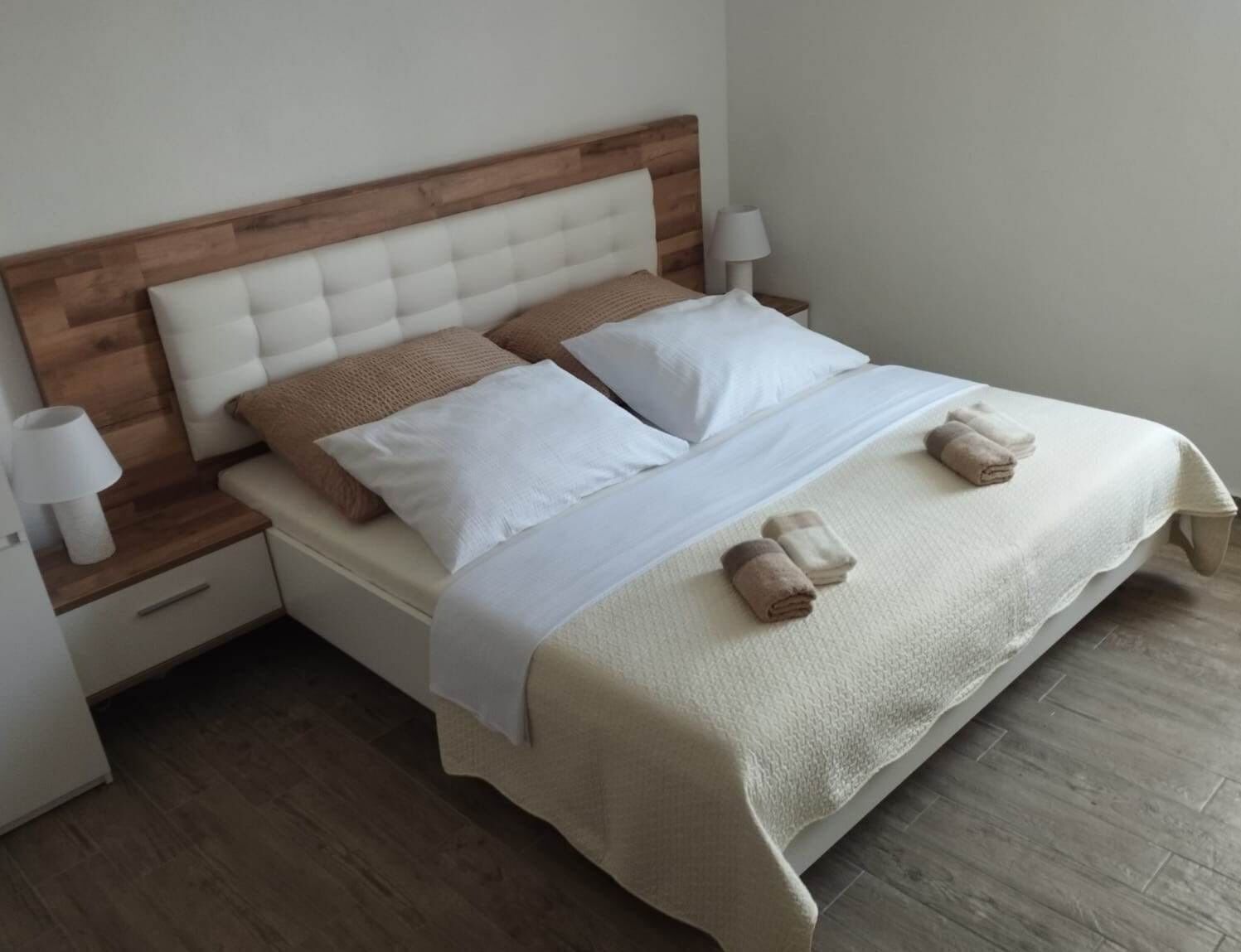 Una cama bien hecha con cabecera de madera, dos lámparas de noche y toallas enrolladas sobre la cama.