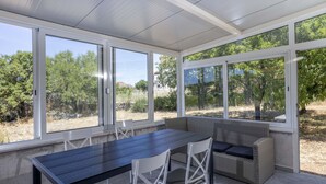 Un porche cubierto con una mesa y sillas de comedor, un sofá y grandes ventanales con vistas a zonas verdes.