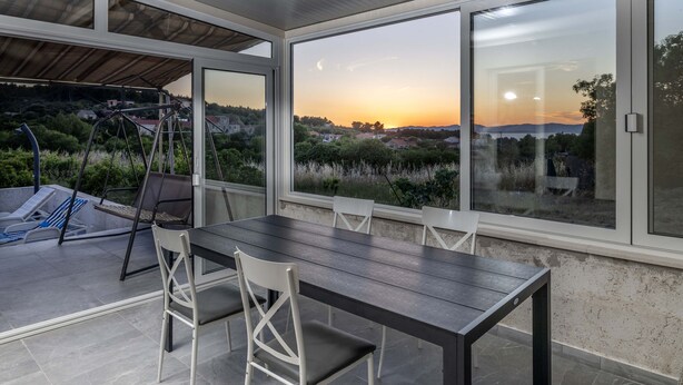 Un comedor con mesa y sillas, ventanas amplias con vista a un atardecer y un paisaje, y un área de estar al aire libre techada con un columpio.