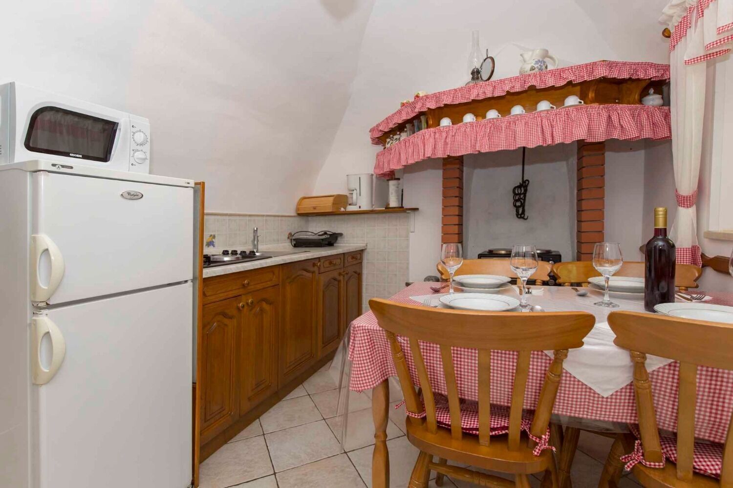 A compact kitchen with a microwave, stove, and sink. A dining table with chairs is set for two, and a wine bottle is on the table.