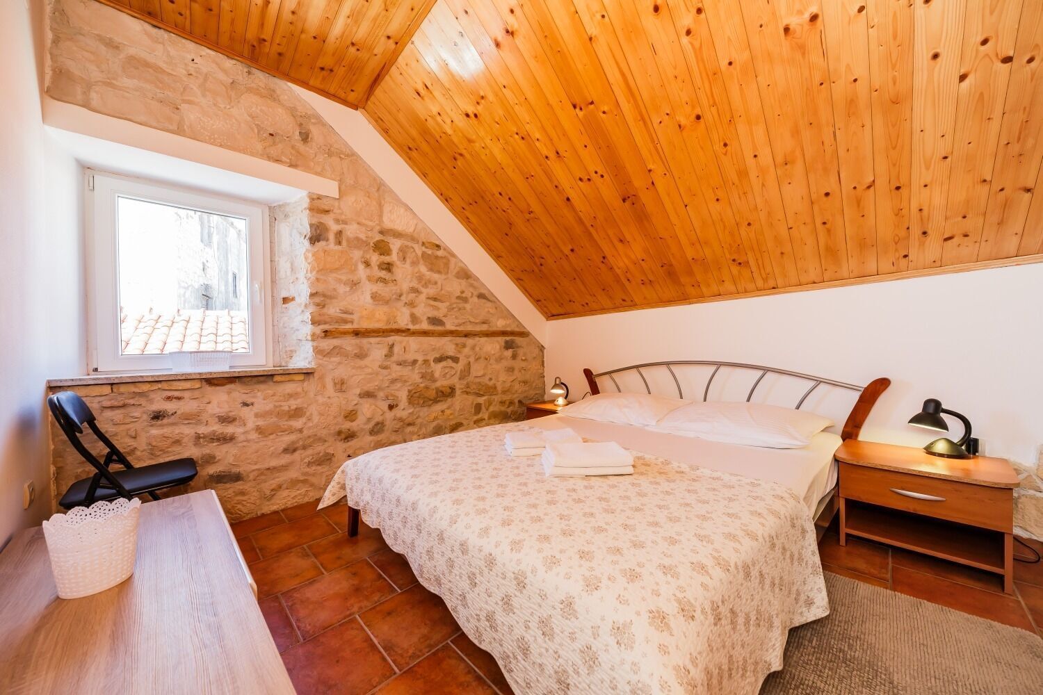 Une chambre avec un mur en pierre, un plafond en bois, un lit recouvert d’une couvre-lit à motifs floraux, une table de chevet avec une lampe et une chaise près de la fenêtre.