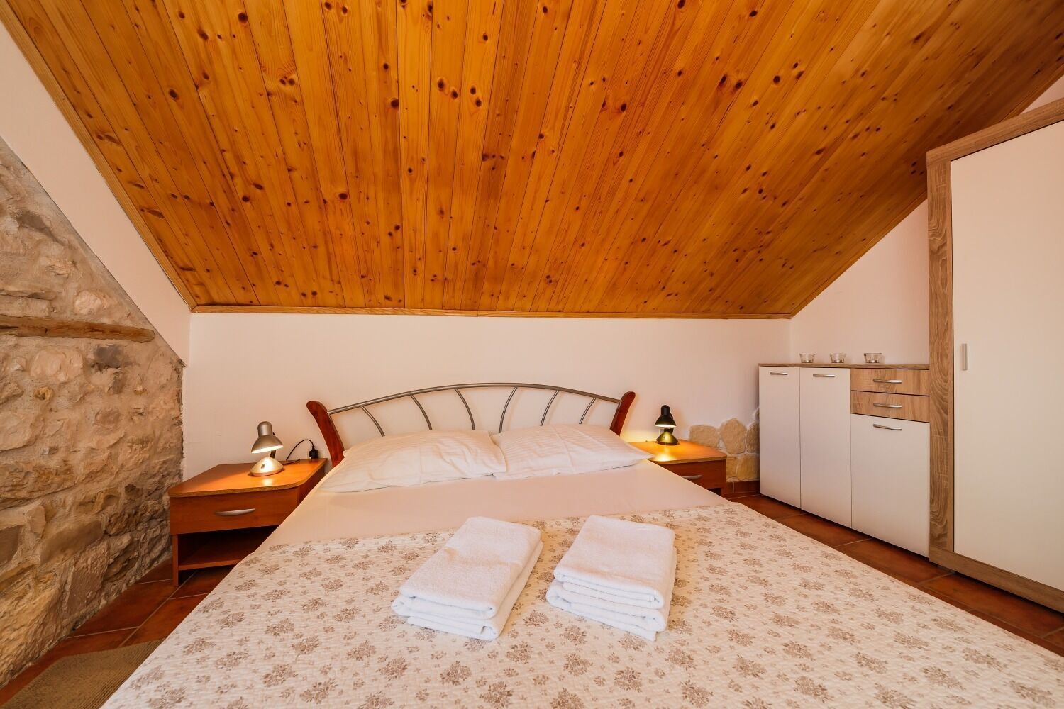 A bedroom with a wooden ceiling, a stone wall, a bed with two white towels, a nightstand with a lamp, and a wardrobe.