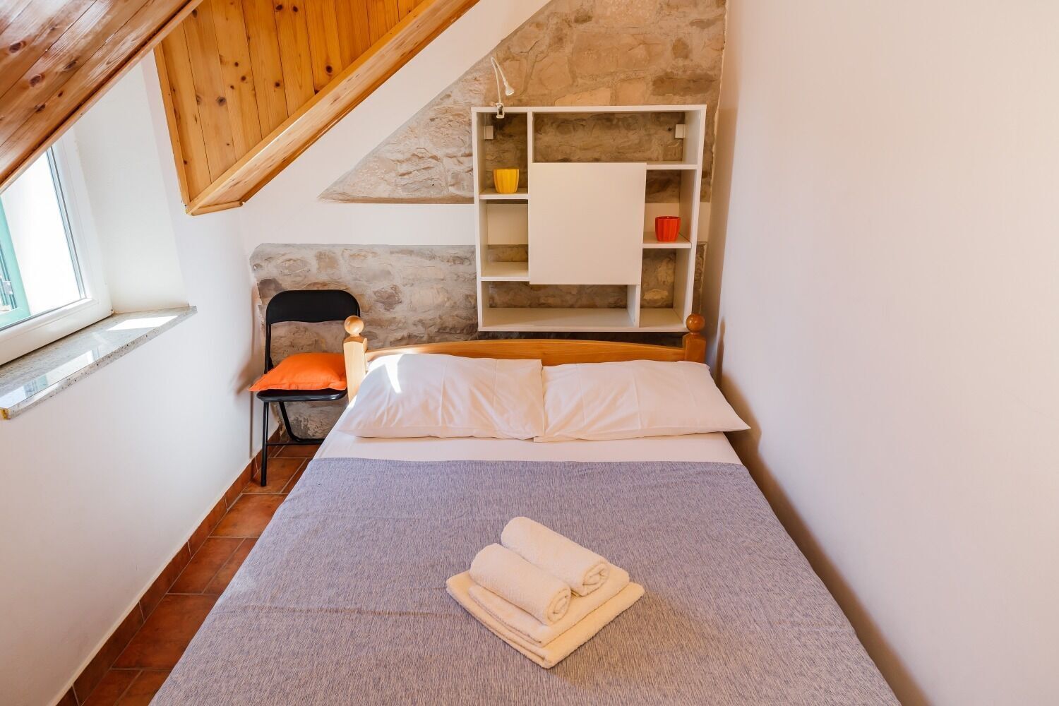 A bedroom with a bed, a chair, a shelf, and a window.