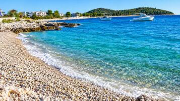 Une plage de galets bordée d’une eau d’un bleu limpide, avec des bateaux et des bâtiments en arrière-plan.